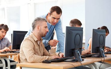 Group of adult students attending computer course. Focus on teacher talking with an adult man.